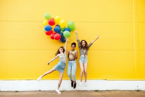 英語圏の国で、黄色い壁に向かってカラフルな風船を持ってジャンプする 3 人の若い女性。