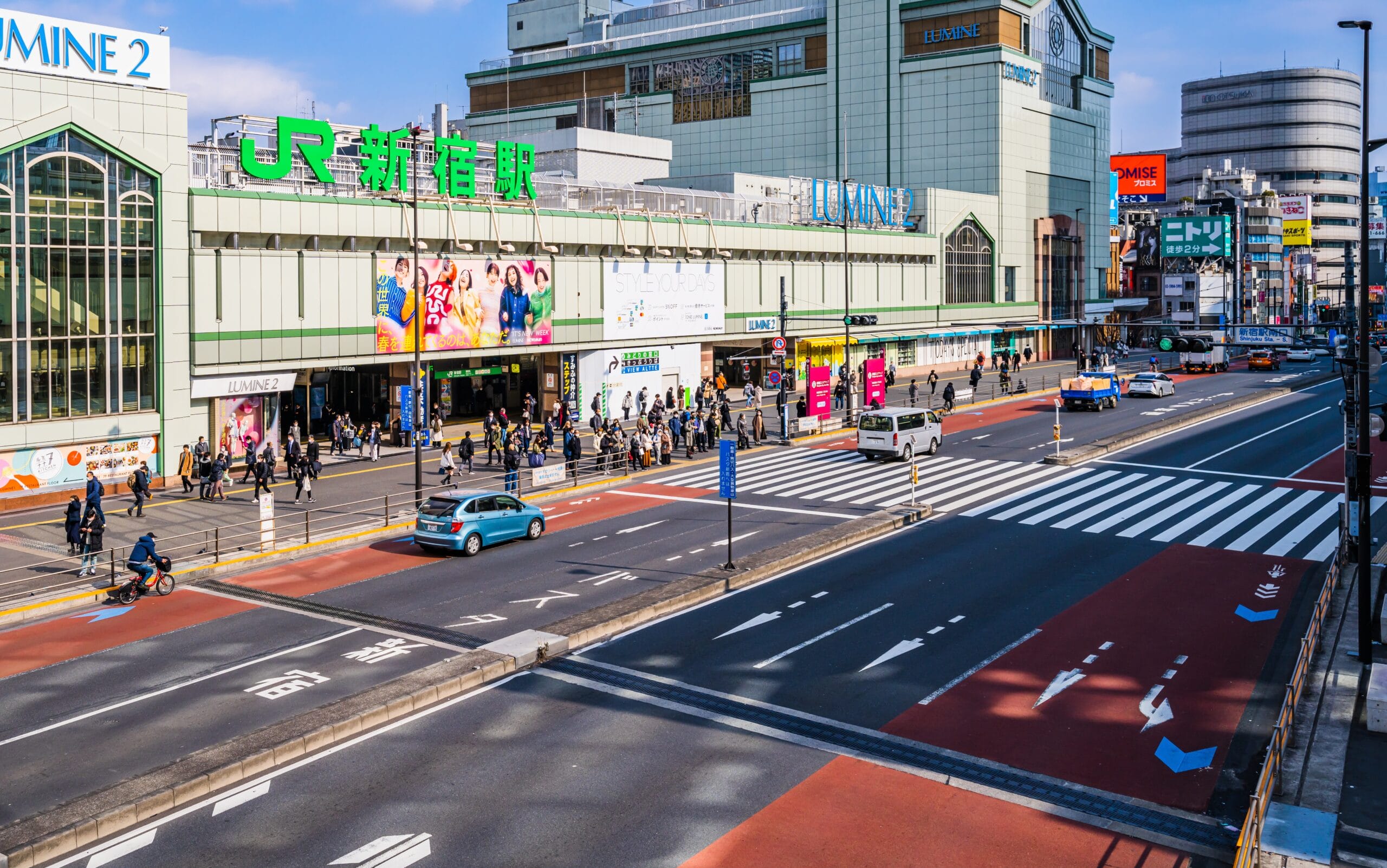 東京の JR 新宿駅前にある繁華街。晴れた日には、高層ビルに囲まれ歩行者と車両が行き交う。活気に満ちた環境は、地元のカフェでの英会話マンツーマンセッションに最適な背景を提供します。.