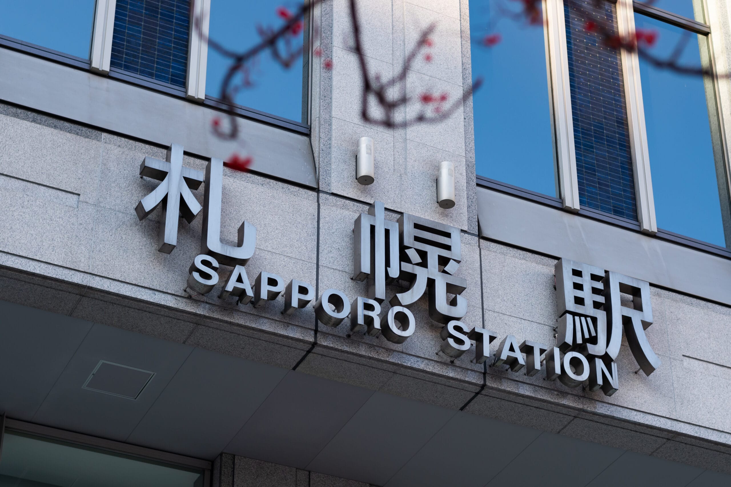 大きな窓のあるグレーの建物にある札幌駅の標識。日本語と英語の表記があり、旅行者にとってのナビゲーションのしやすさを強調しています。札幌で英会話マンツーマンを目指す方に最適な立地です。.