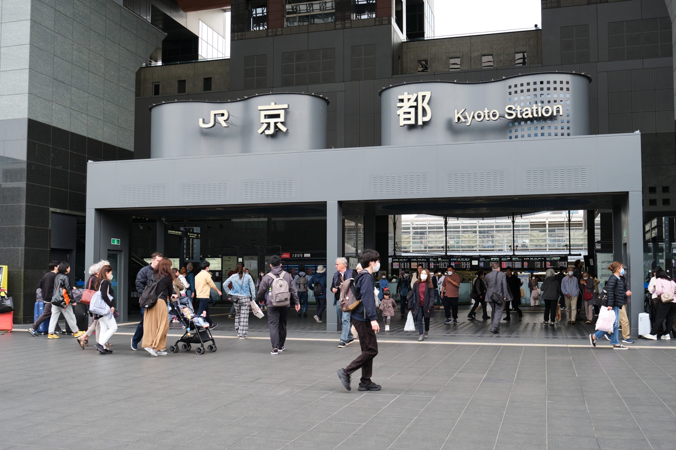 「JR」と「京都駅」の看板が掲げられた京都駅の入り口前は人々が行き交い、英会話マンツーマンの交流や、活気に満ちた街並みに浸るのに最適な賑やかな雰囲気を作り出しています。.