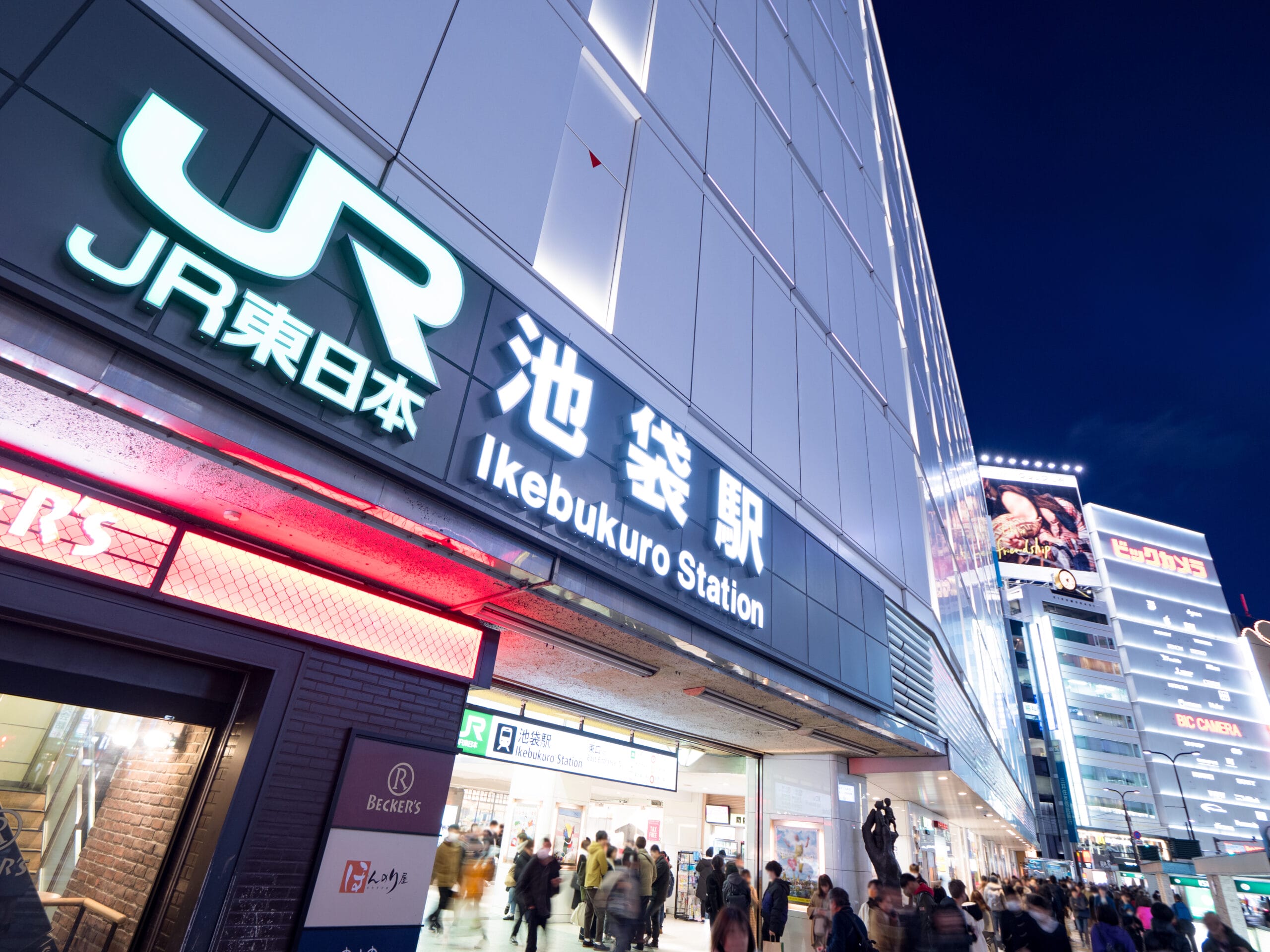 夜の池袋駅入口は、明るい看板の光の下、英会話マンツーマンなどのイベントを暗示する電飾看板が背景にあり、多くの人で賑わっています。.