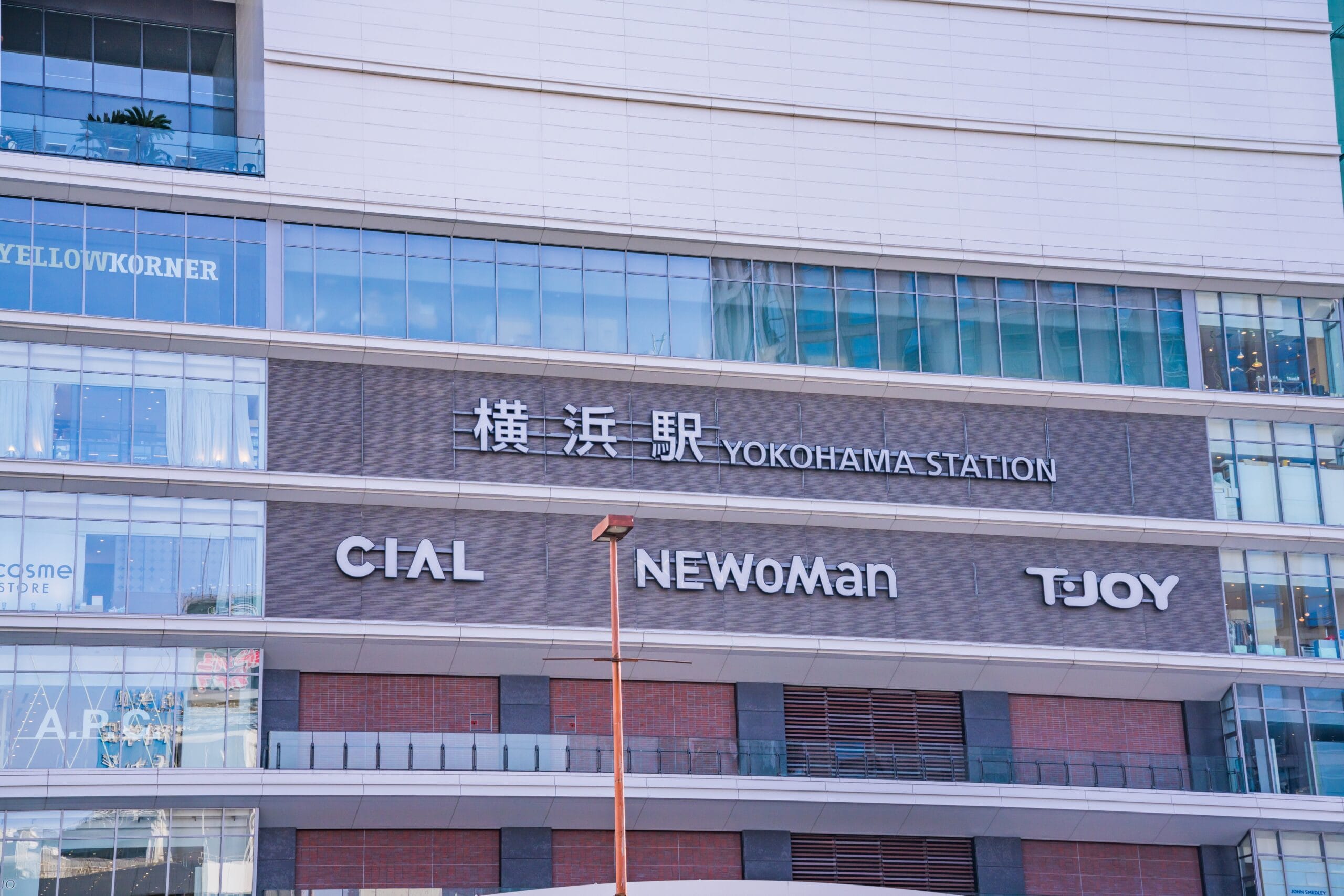 この画像は日本の横浜駅の外観を示しており、高層ビルのファサードに対してCIAL、NEWoMan、TJOYの看板が強調されています。活気あふれる横浜の街にあるこの賑やかなエリアは、文化的な楽しみを探索したり、近くで英会話マンツーマンのレッスンを見つけたりするのに理想的な場所です。.