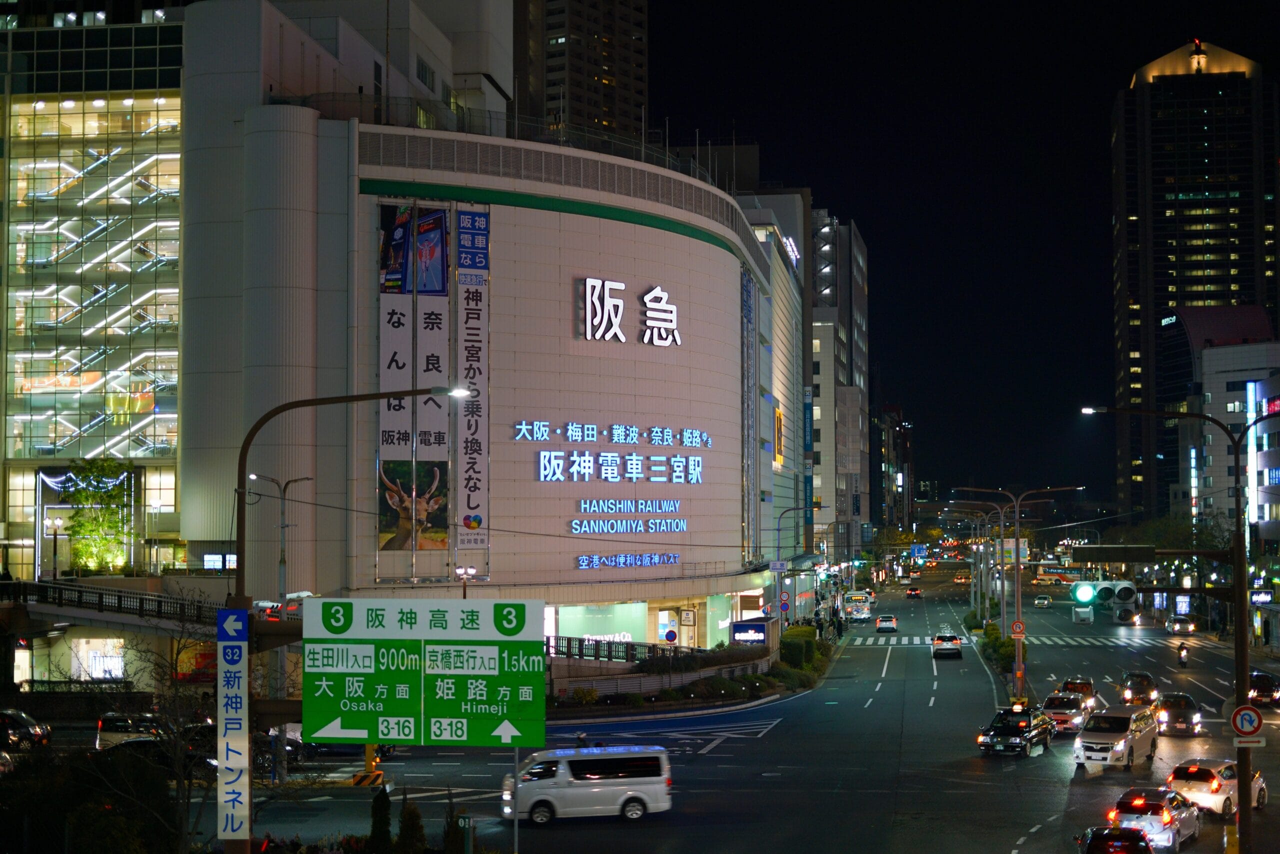 夜の賑やかな街路には、阪急電鉄の電飾看板や日本語の道路標識が灯り、街を明るく照らしています。高層ビルの間を車が行き交い、神戸三宮の看板が目立つように立ち、活気に満ちた都会の風景の中を旅人を案内しています。.