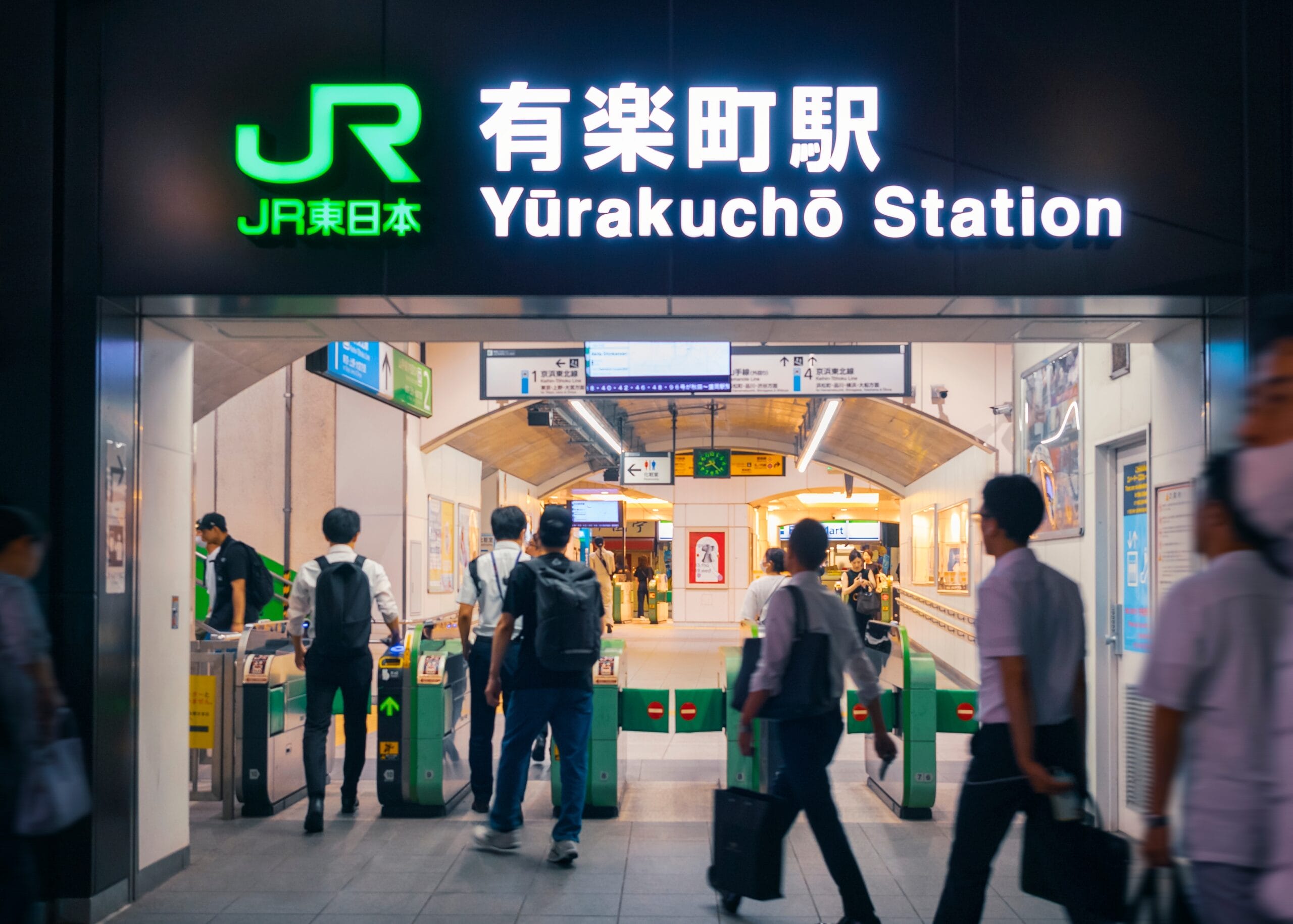 有楽町駅の入口は改札を通る人々で賑わっています。頭上には日本語と英語の鮮やかなネオンサインが光ります。駅構内の駅員の近くには、英会話スクールマンツーマンの広告が通勤客にマンツーマンで語学力の向上を呼びかけています。.