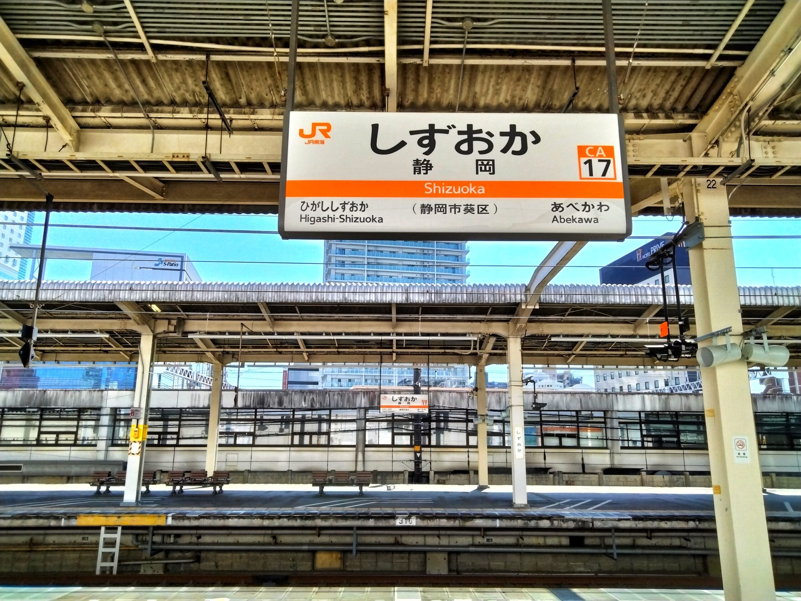 静岡県の静かな駅のホームには、「掛川・静岡」などの行先名が日本語と英語で書かれた標識が設置されています。背景には都市の建物が見え、英会話スクールマンツーマンが言語スキルを向上させる可能性のある都市を反映しています。.