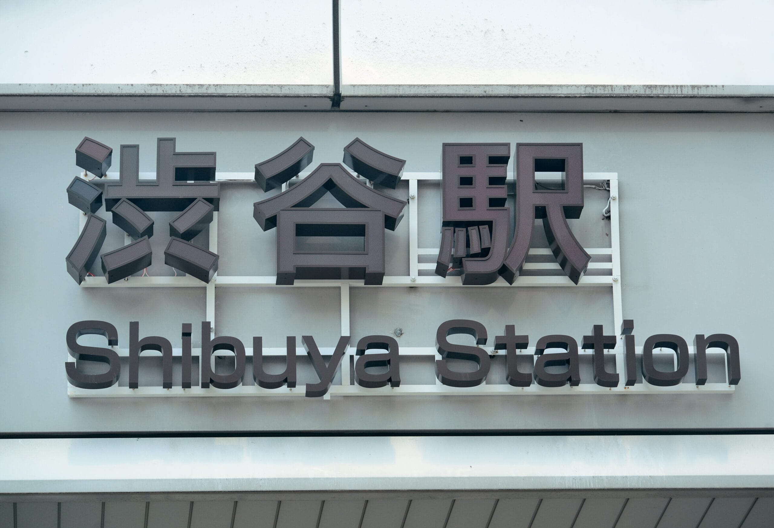 渋谷の建物の外壁に設置された英語と日本語の文字で「Shibuya Station」と書かれた看板は、活気に満ちた文化の融合を反映しており、近くのにぎやかな英会話スクールマンツーマンとは魅力的な対照を成しています。.