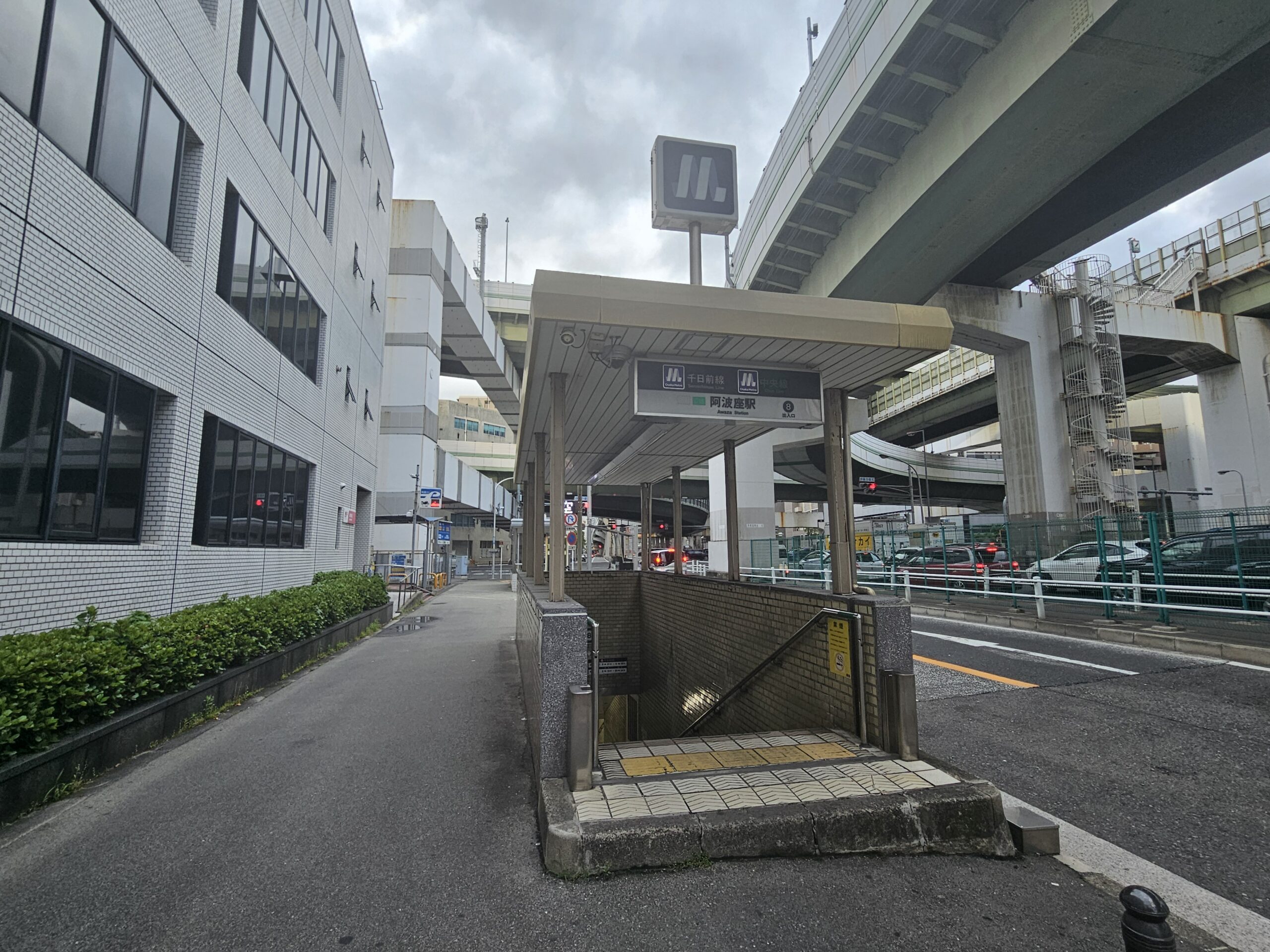阿波座近くの通り沿いにある地下鉄駅の入り口。曇りの空の下、背景には高層ビルや高架道路、そしてすぐそばにある英語教室が見える。.