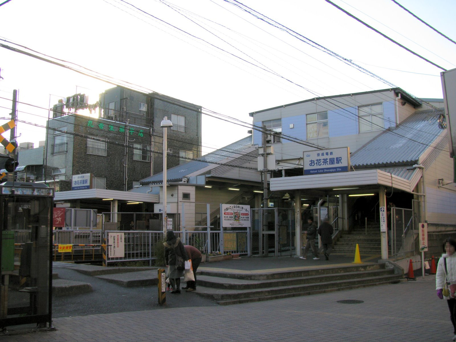 日本語の看板が掲げられた小さな駅に、人々が出入りしている。「お花茶屋」と書かれた駅舎は傾斜した屋根をしており、頭上には電線が走っている。その近くでは、「英会話教室」という看板が英語レッスンの宣伝をしている。.
